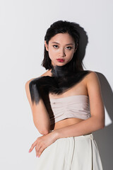 young asian woman with bare shoulders and hand in black paint looking at camera on white.