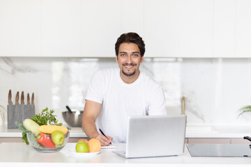 Fototapeta na wymiar Young handsome and fit man sitting at the table in the kitchen and writing his diet plan in notebook. A lot of fruits and vegetables which on the table also included in his ration