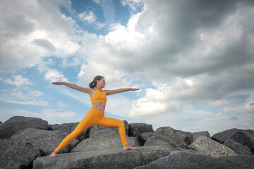 Fit, sporty woman practicing yoga and exercising on rocks outside, warrior pose.