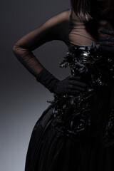 Naklejka premium cropped view of woman in black gothic dress and gloves posing with hand on hip on dark grey.