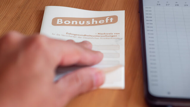 Bonusheft zur zahn&auml;rztlichen Vorsorgeuntersuchung | Kalender auf Smartphone | Zahnarzttermin vereinbaren | Hand bl&auml;ttert Bonusheft auf