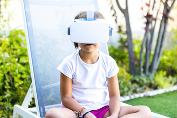 Little girl with virtual reality glasses, sitting on a sun lounger in the garden of her house. Metaverse, VR, game, digital and simulation concept.