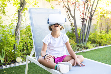 Little girl wearing virtual reality goggles, looking up, sitting on a deckchair in her garden at...