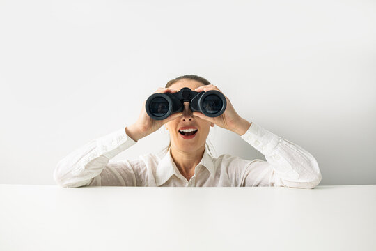 Surprised young woman in shirt looking through binoculars peeking out from behind desk