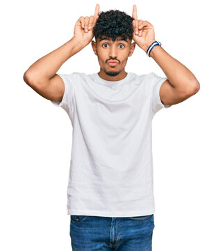 Young arab man wearing casual white t shirt doing funny gesture with finger over head as bull horns
