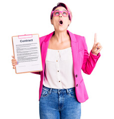 Obraz na płótnie Canvas Young beautiful woman with pink hair holding clipboard with contract document amazed and surprised looking up and pointing with fingers and raised arms.