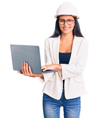 Young beautiful latin girl wearing architect hardhat holding laptop looking positive and happy standing and smiling with a confident smile showing teeth