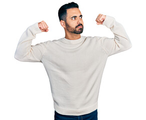 Young hispanic man with beard wearing casual white sweater showing arms muscles smiling proud. fitness concept.