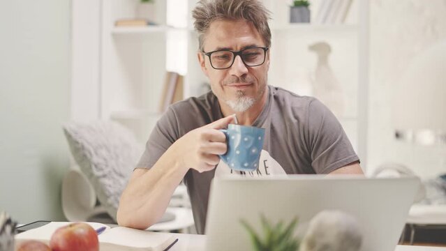 Man sitting at desk at desk drinking morning coffee and reading news feed on laptop computer. Middle aged, mid adult, mature age man working in home office