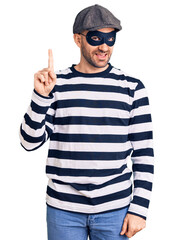 Young handsome man wearing burglar mask pointing finger up with successful idea. exited and happy. number one.