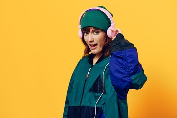 a happy, joyful woman in a green jacket and hat enjoys music standing in pink headphones on a yellow background holding them with her hands and looking at the camera