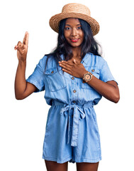 Young indian girl wearing summer hat smiling swearing with hand on chest and fingers up, making a...