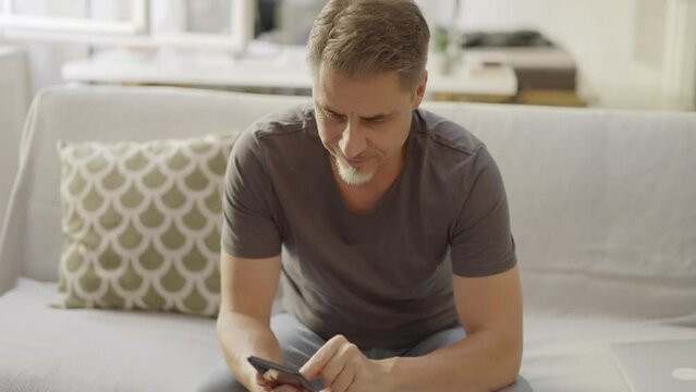 Older man at home, sitting on couch in living room, using phone, checking social media. Portrait of happy, mature age, middle age, mid adult caucasian man in 50s, smiling.