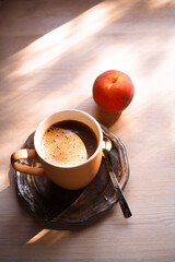 A cup of coffee on vintage matal plate on white wooden table with a peach at morning light, top view, summer morning concept.