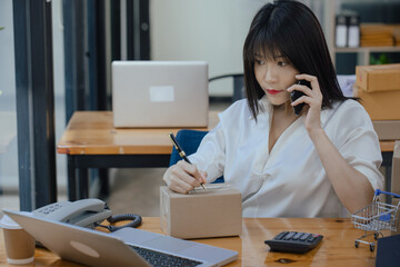 Asian businesswoman using smartphone with parcel boxes and checking orders sitting at office desk.  Business online selling and online shopping concepts.