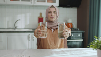 Muslim hijab dressed woman in hijab holding a dirty glass in one hand and a clean glass in the...