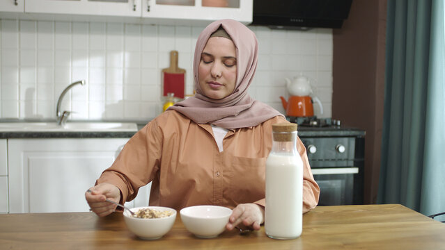 Happy Hijab-dressed Muslim Sitting At The Kitchen Table At Home Pouring Milk Into A Bowl Of Granola With Flakes And Having Breakfast. A Woman Dressed In A Hijab Is Eating Cereal In The Kitchen.