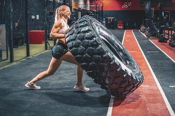 Fit blonde woman flipping heavy tire at gym.