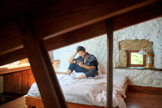 Stylish Hispanic man chatting on smartphone on bed