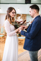 The bride in a white lace dress, glasses, with feathers in her hair holds a boho style wedding bouquet and puts a wedding ring on the groom's finger. Wedding ceremony in the registry office.