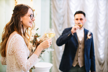 The bride in a lace dress with feathers in her hair holds a glass of champagne and smiles. In the background, the groom drinks champagne and looks at the girl. Wedding ceremony.