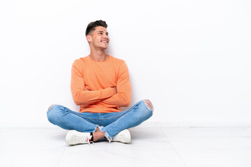 Young man sitting on the floor isolated on white background looking up while smiling