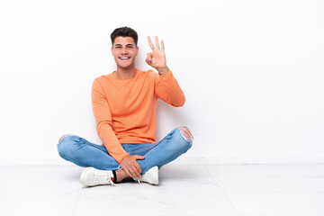 Young man sitting on the floor isolated on white background happy and counting three with fingers