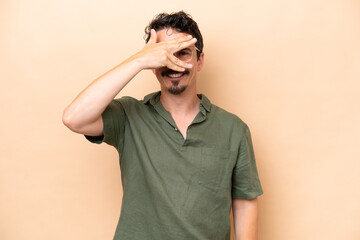 Young caucasian man isolated on beige background covering eyes by hands and smiling
