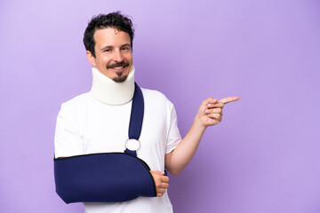 Young caucasian man wearing a sling and neck brace isolated on purple background pointing finger to...