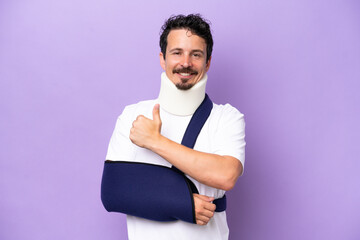 Young caucasian man wearing a sling and neck brace isolated on purple background giving a thumbs up gesture