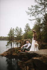 The bride and groom are sitting in a stone canyon by the lake. The groom is sitting on a rock. The bride in a white dress, leather jacket, and hat holds a wedding bouquet and looks at the lake.