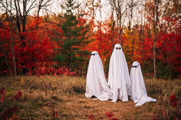 Three children dressed as ghosts wearing sunglasses play in front of colorful autumn foliage