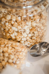 A large glass container of popcorn with a metal scoop at a wedding for a late night snack