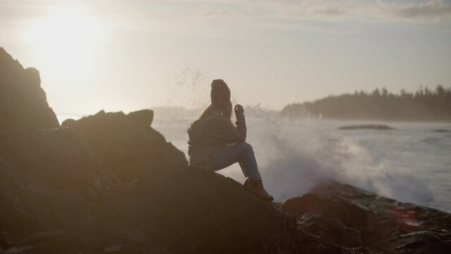 Women sits on rocks by the ocean and reflects as waves crash behind her during sunset
