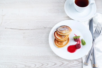 On a light background, pancakes with powdered sugar and raspberries, cutlery and a cup of tea.