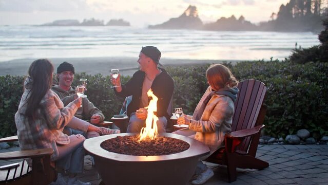 Joyful young friends sitting around fire cheersing glasses at sunset, ocean views
