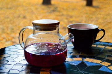 A teapot with hot berry red tea on the table against the background of fallen yellow leaves. Cup in the background. selective focus. Autumn mood, yellow leaves, bright autumn. Postcard, background