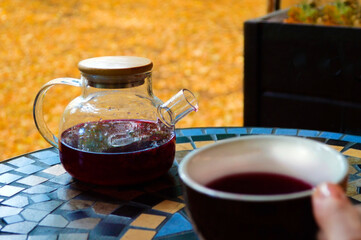 Autumn, a teapot with hot berry tea on the table against the background of fallen leaves. The cup in the girl's hand is out of focus. Autumn mood, yellow leaves, bright autumn. Postcard, background