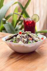Eggplant salad, parmesan cheese, olives and lettuce served in a white bowl on a wooden table in a restaurant. Accompanied by a glass of red juice.