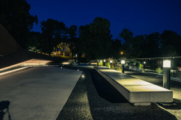 Summer night city park. Wooden benches, street lights, and green trees. The tiled road in the night park with lanterns. Illumination of a park road with lanterns at night. Lviv