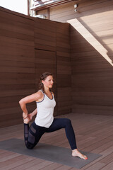 Naklejka premium Beautiful strong muscular woman doing stretching on mat on terrace outdoors. Healthy young adult girl in sportswear stretch alone. Fit thin gimnast doing yoga, training on mat