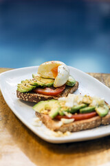Avocado, tomato and egg toast, served on a white plate on a wooden table. Healthy food, rich and nutritious breakfast.