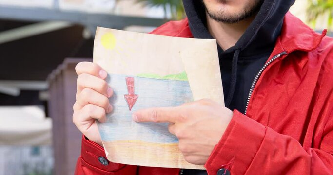Person in a hoodie unfolds a drawing on which arrow indicates place in the sea, where something valuable is located, front view. Person points to this place with finger