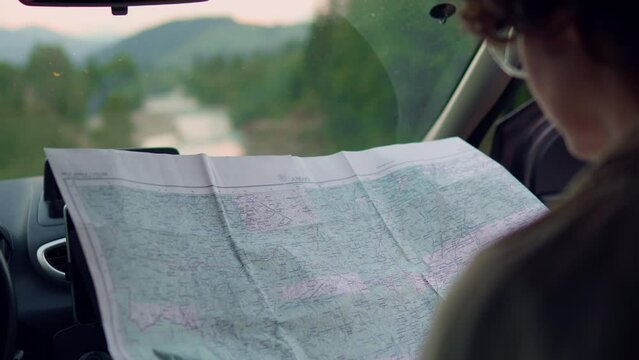 Traveler girl sits in the car looks at the map. A young active girl engaged in hiking came to the place building plans a route while sitting in a car against the backdrop of mountains
