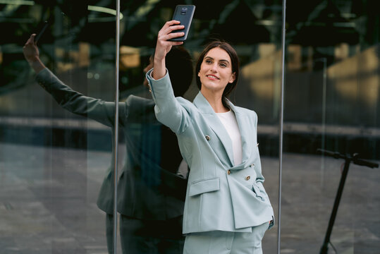Delighted businesswoman in suit taking selfie near glass building - Powered by Adobe