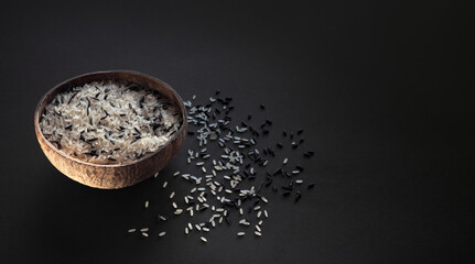 Wild rice in a coconut bowl with spilled rice grains on a table for vegan menu design in minimalism style. Mix of the rice cereals on black background in natural sun light with copy space.