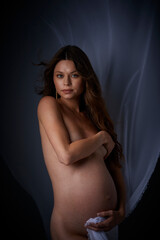Naklejka premium Beautiful young pregnant serious sexy woman with long curly hair and big belly, without clothes sitting in dark studio and covers her breasts with her hand