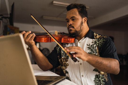 Ethnic man playing violin near table with laptop in house