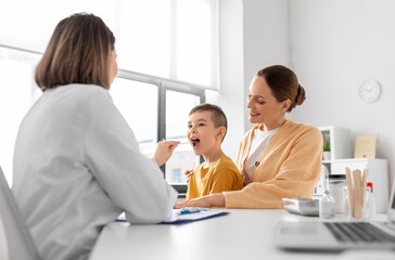 Obraz premium medicine, healthcare and pediatry concept - smiling mother with little son and doctor with tongue depressor checking patient's throat at clinic