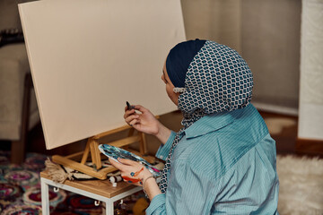 A woman in a hijab paints on canvas with a brush and tempera